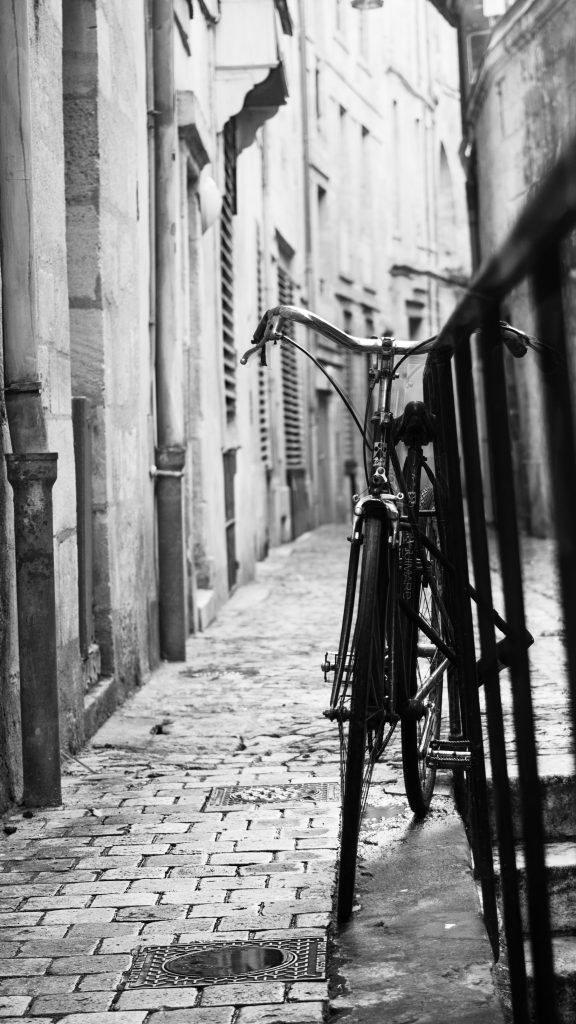 Photographie en noir et blanc d'un vélo accroché à une rambarde. Le vélo est situé sur le tiers droit de la photo, et la rambarde continue au delà du cliché sur le même côté. La mise au point est faite sur le vélo, laissant le reste du cliché légèrement flou.

Le reste de la photo montre une petite ruelle dont les murs en pierre ne sont pas tout à fait droits. La route est pavée et certains reflets sur les pierres indique qu'elles sont mouillées.