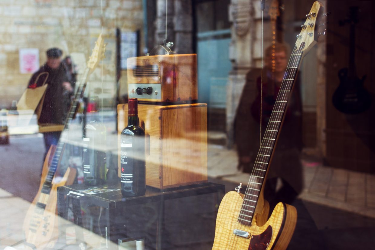 Photographie en couleur d'une vitrine d'un magasin qui expose des bouteilles de vin et des instruments de musiques. Sont visibles en premier plan et sur la gauche de l'image deux guitares. Au centre de l'image un ampli posé sur une caisse en bois. Deux bouteille de vins sont posées devant elle.