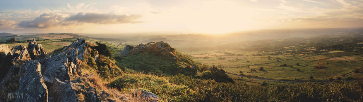 Photo panoramique au sommet des Monts d'Arrée en Bretagne. Sur la partie gauche on observe des rochers sur lesquels des herbes folles poussent. Sur la partie droite, en contrebas, une route serpente au milieu de grandes étendues d'herbes. Le soleil se couche sur l'horizon, dans un ciel légèrement nuageux.