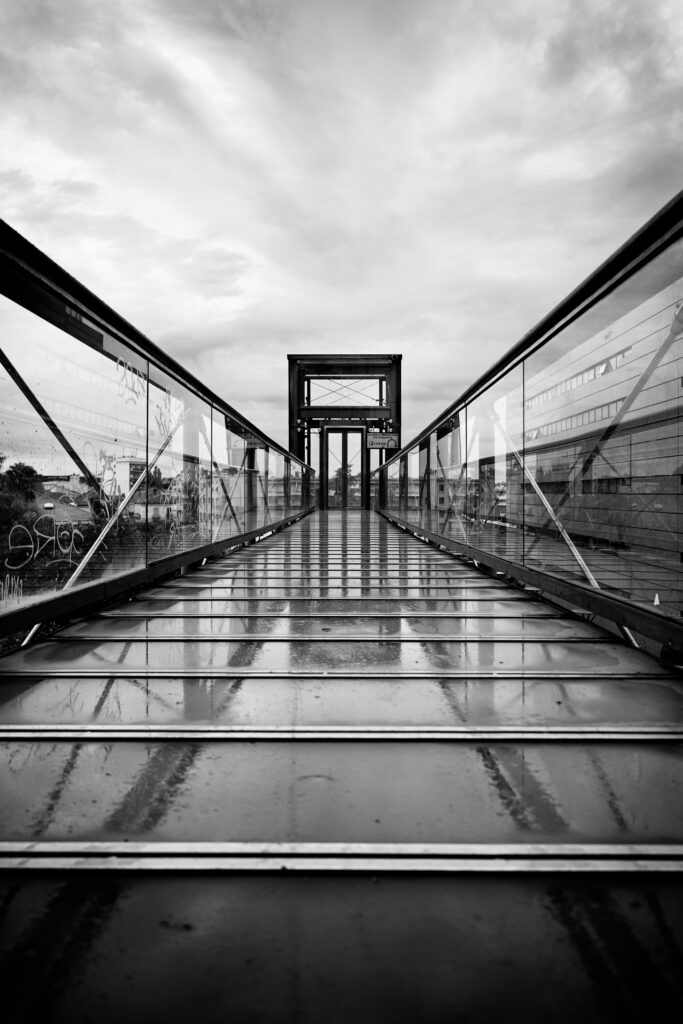 Photographie en noir et blanc d'un ascenseur quasiment entièrement fait de verre. Le chemin qui y mène est en verre et reflète tant le ciel que l'ascenseur. Les rambardes laissent apercevoir à gauche des immeubles ainsi que de la végétation, tandis qu'à droite un grand bâtiment contemporain est visible. Enfin, dans la partie haute de la photo, c'est un ciel nuageux qui surplombe la scène.