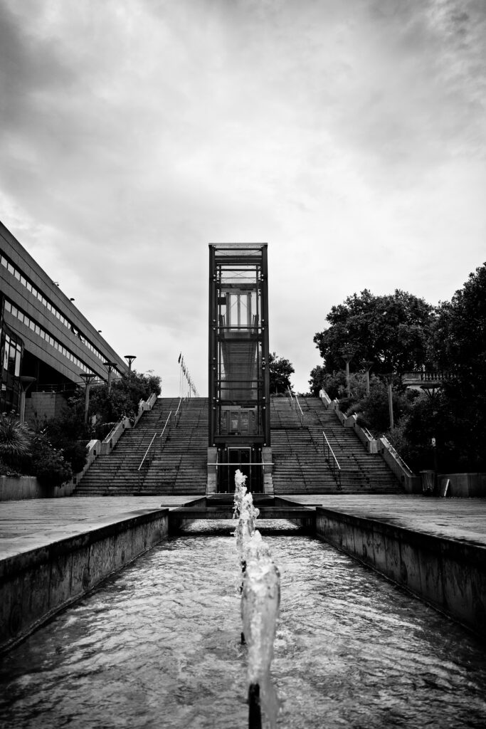Photographie en noir et blanc dont le sujet principal est un ascenseur.

Au premier plan plusieurs jets d'eau sont visible, dans l'alignement avec l'ascenseur. Ils font parti d'un chemin d'eau qui court tout le long de la promenade. 

Au second tiers, au centre, un grand escalier, bordé à droite de végétation diverses, et à gauche d'un bâtiment moderne. Séparant l'escalier en deux se trouve l'ascenseur.

Enfin au tiers supérieur, un ciel gris et nuageux est visible.