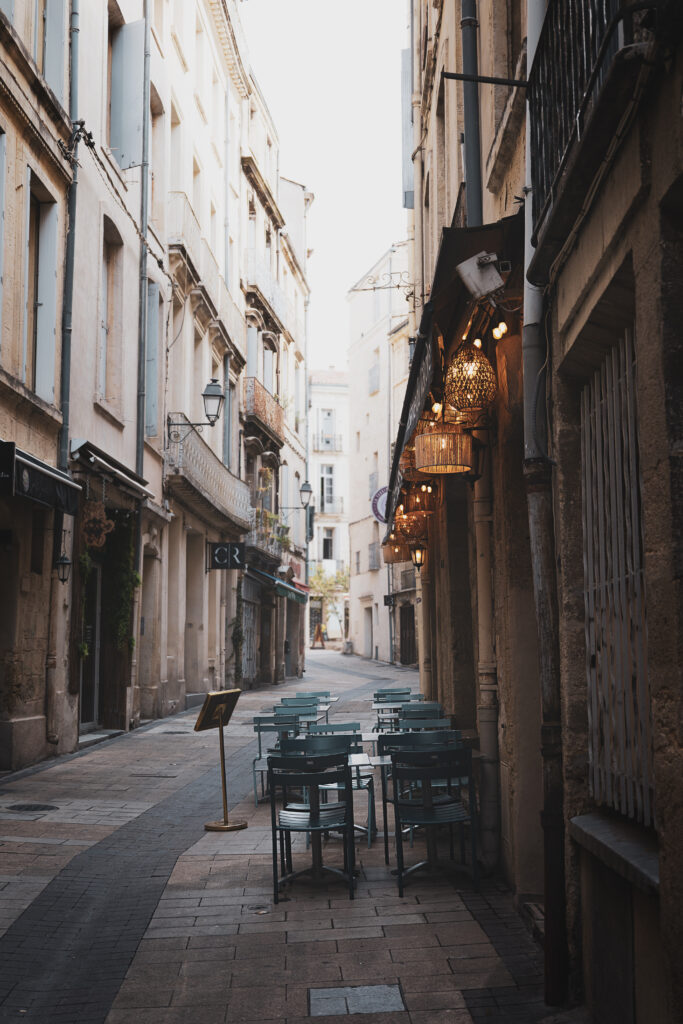 Photographie montrant une rue ancienne, bordée d'immeubles haussmanniens. La mise au point est faite sur des lampions accrochés sous le volet déroulant d'un restaurant. En dessous, plusieurs chaises et tables attendent les clients.
