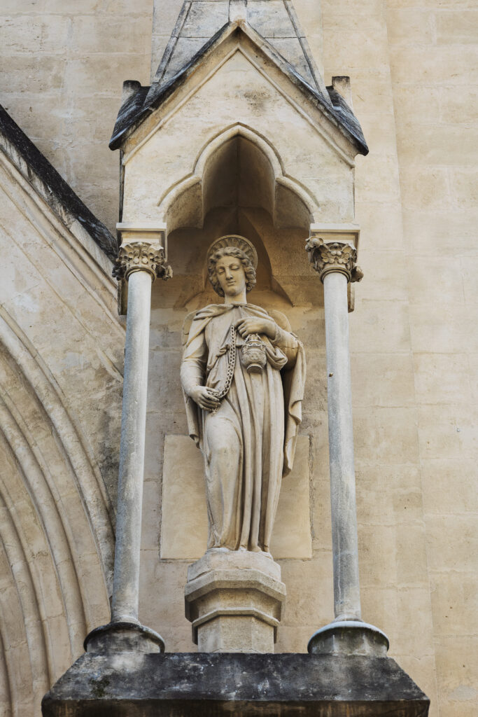 Photographie d'une statue d'ange sur la devanture d'une église, protégée par une voute décorée, tenue par deux piliers.
L'ange, tenant dans sa main gauche un encensoir. Sa main droite soutien la chaine de l'encensoir au niveau de sa hanche.