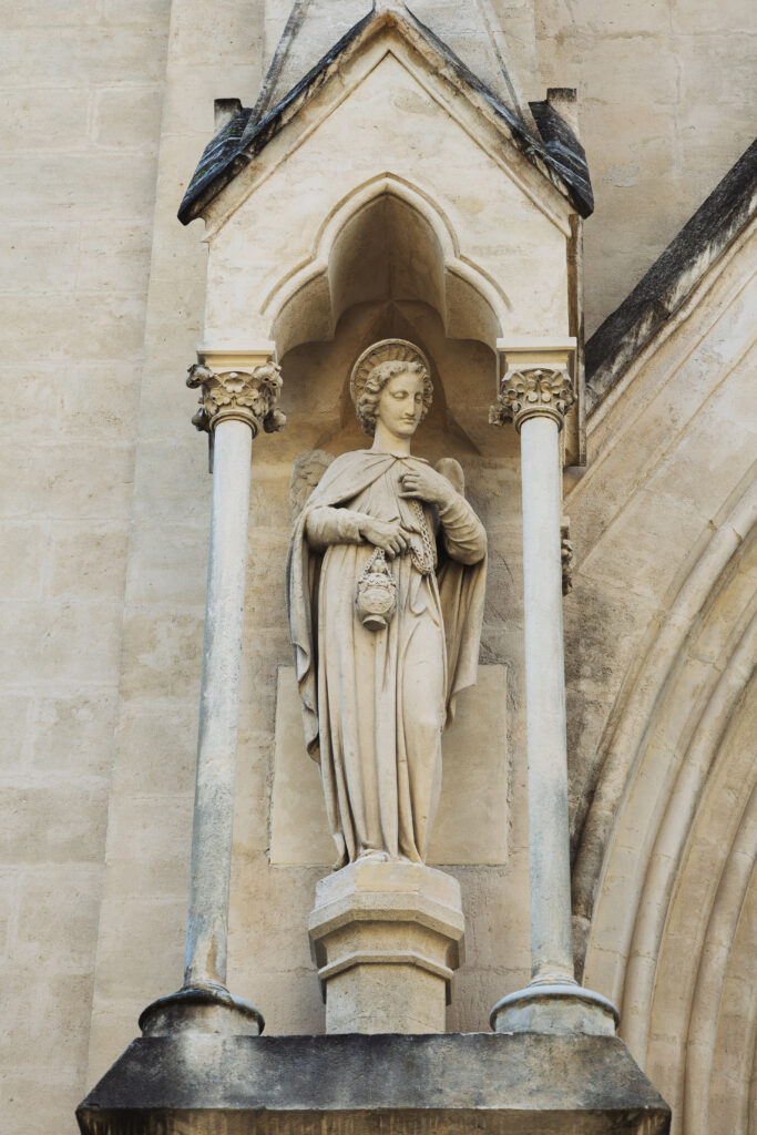 Photographie d'une statue d'ange sur la devanture d'une église, protégée par une voute décorée, tenue par deux piliers.
L'ange, tenant dans sa main droite un encensoir. Sa main gauche repose sur sa poitrine.