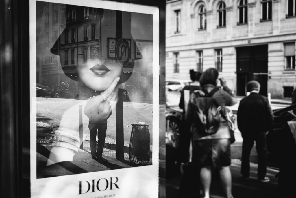 Street photographie d'un abri bus. La mise au point est faite à gauche, sur l'affiche publicitaire Dior, d'une femme avec un chapeau cachant ses yeux, la main soutenant son visage. Dans le reflet de la vitre on voit une personne attendre.
A droite, plusieurs personnes attendent également.