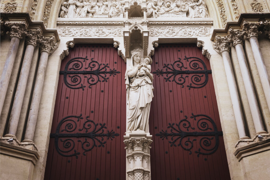 Photographie d'une façade d'église. On y voir deux portes rouge entourant une statue d'une vierge à l'enfant. Les portes sont décorées d'ornement en fer forgé noir. Autour de nombreuses sculptures, tant au niveau des piliers ouvragés que sur le fronton, qui semble narrer la mise au tombeau.