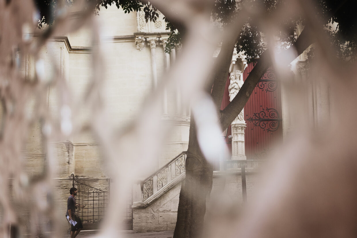 Photographie d'un street art fait de morceaux de miroirs. La mise au point est faite sur le reflet, qui montre l'entrée d'une cathédrale, avec deux portes rouges, entourant une madonne à l'enfant. On y voit également la rampe d'un escalier menant à ses portes. En bas à gauche, le reflet d'une personne marchant.