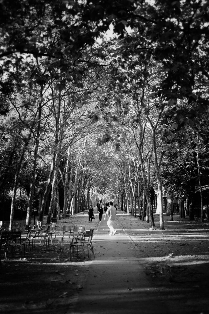 Photographie en noir et blanc d'une allée bordée d'arbres. Au milieu un chemin droit file vers le centre de la photo. Plusieurs personnes marchent sur ce chemin. A gauche plusieurs chaises et tables vides attendent les promeneurs fatigués en quête d'un instant de repos.
