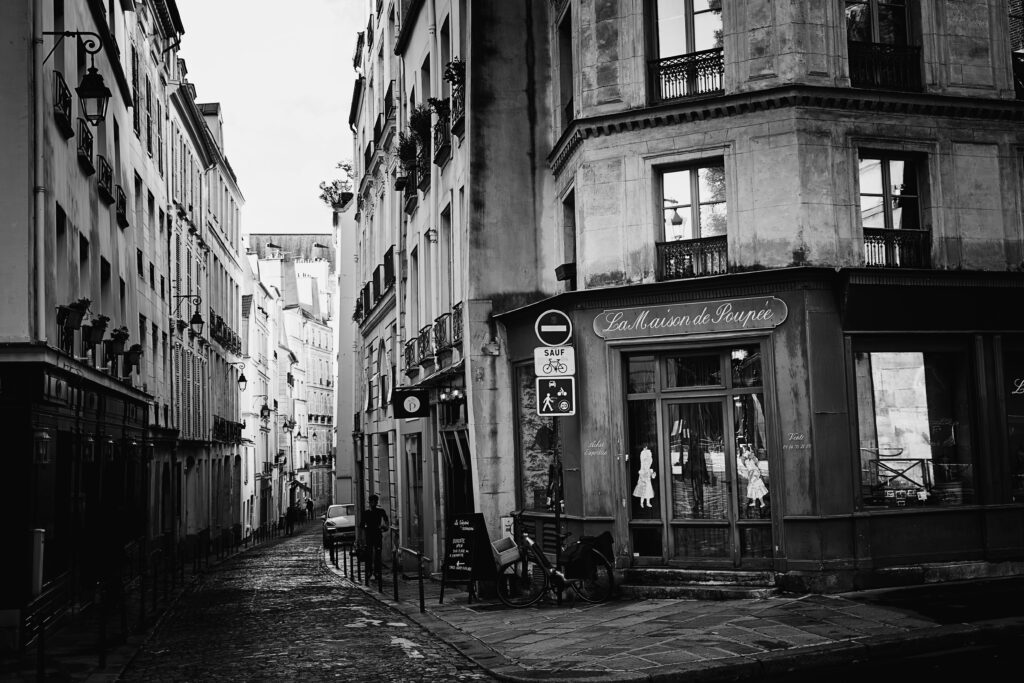 Street photographie d'une rue étroite, au carrefour de plusieurs voies. Sur la droite la mise au point est faite sur la devanture d'un magasin "La maison de poupée". Des poupées sont visibles, affichée sur les vitres entourant la porte d'entrée. 
Un peu plus bas dans la rue adjacente, un piéton est en train de remonter la rue.