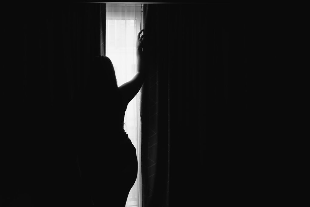 Photographie d'une personne en contrejour. Des rideaux quasiment fermé laisse entrevoir une bande de lumière dans laquelle se découpe une silhouette féminine qui lève le bras comme pour ouvrir le rideau.