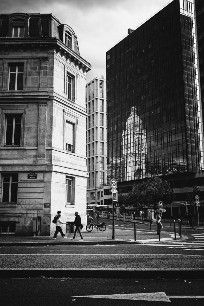 Photographie de rue. On y voit plusieurs batiment, à gauche un immeuble parisien typique du centre ville, en pierre, avec quatre étage dont un sous les toits. De l'autre côté, un batiment ultra moderne, dont le côté exposé est entièrement en verre, reflétant la tour de l'horloge de la gare de Lyon.
Dans le tiers bas, la route et plusieurs personnes marchant.