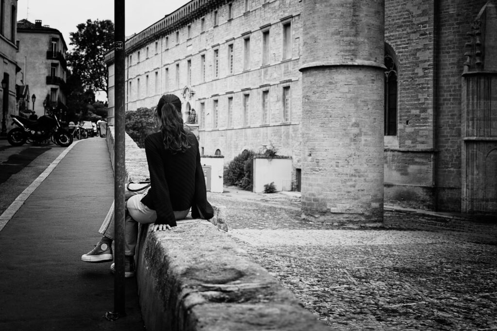 Photographie d'une personne assise sur un muret, semblant attendre quelqu'un. Elle porte un pull sombre et un pantalon clair. Ses longs cheveux sont ramenés en queue de cheval.

En arrière plan un batiment en pierre abritant une faculté de médecine. A droite on observe un immense pilier également en pierre et le début d'une arche, c'est l'entrée d'une cathédrale.