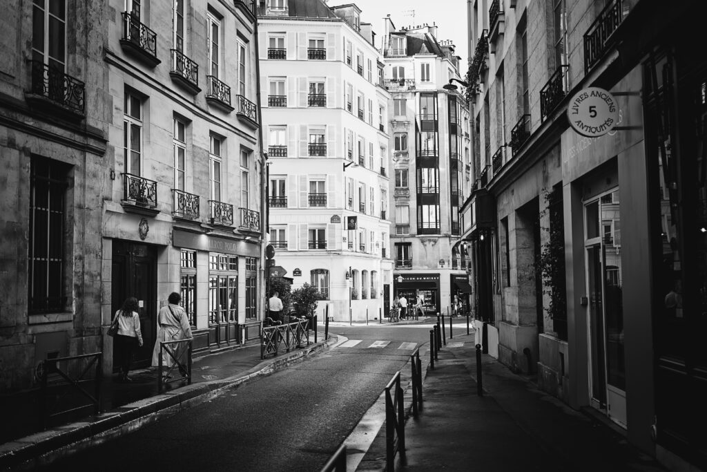 Photographie de rue à Paris, montrant plusieurs bâtiments avec au centre de la photo un carrefour. A gauche une librairie avec une enseigne notée 'Livres ancien antiquités" et un cinq au milieu. A droite deux personnes se promènent sur le trottoir. 