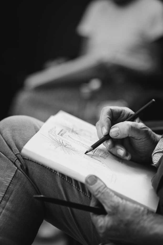 Photographie en noir et blanc d'une personne en train de dessiner. On y voit un carnet, que la personne tient sur sa cuisse avec l'une de ses mains. L'autre est en train de tenir un crayon. Dans le fond on observe une autre personne en train de dessiner.