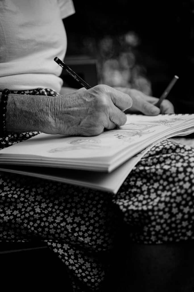 Photographie en noir et blanc d'une personne en train de dessiner. Elle tient son carnet sur ses cuisses, recouverte d'une jupe avec un motif de très petites fleurs blanche. Ses deux mains tiennent un crayon, l'une dessine, pendant que l'autre retient le carnet.