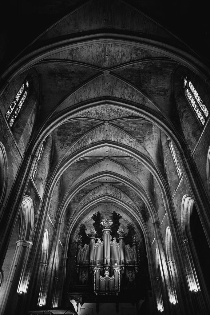 Photographie en noir et blanc de l'intérieur de la cathédrale Saint Pierre à Montpellier. L'accent est mis sur les voutes du plafond. Au centre du tiers bas de la photographie se trouve l'orgue, dont l'éclairage projette les ombres sur la dernière voute derrière lui.
