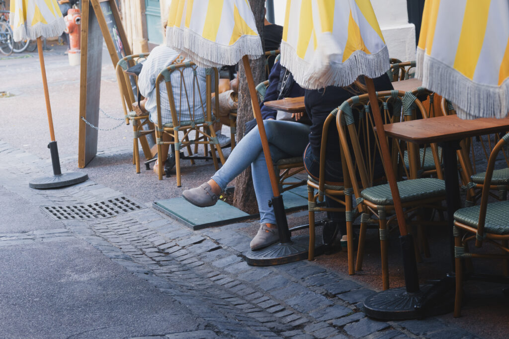 Photographie de rue, d'une terrasse de café. On y voit plusieurs tables et chaises. Vers le milieu, on voit deux personnes attablées, l'une d'entre elle a les jambes croisées en direction de la rue. Quatre parasols fermés sont disposés, dont un cache la tête des personnes.