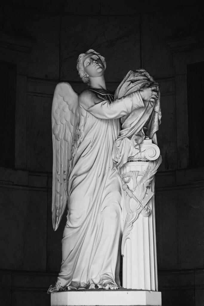 Photographie en noir et blanc d'un ange, debout à l'abri dans une coupole ouverte, regardant vers le haut et drapant une urne d'un linceul.