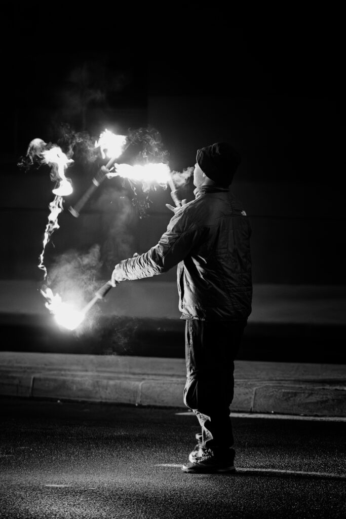 Photographie de rue en noir et blanc montrant un jongleur de feu, au milieu d'une route, de trois quart dos, en train de faire virevolter trois torches allumées.