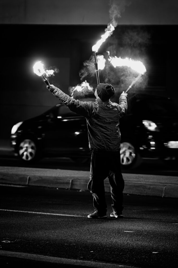 Photographie en noir et blanc d'un jongleur de feu. Il tient dans chacune de ses mains une torche, pendant qu'une troisième est en équilibre sur sa tête. Il se trouve de trois quart dos. Dernière lui une voiture passe.