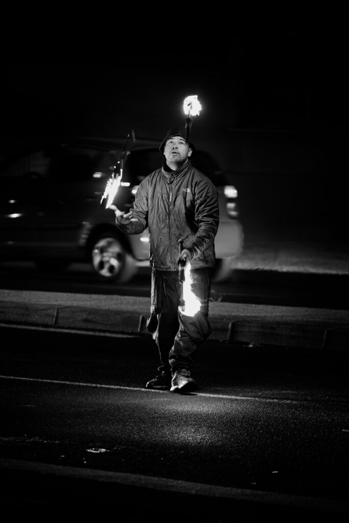 Photographie de rue en noir et blanc montrant un jongleur de feu, au milieu d'une route, de face, en train de faire virevolter trois torches allumées. Derrière lui une voiture est en train de passer.