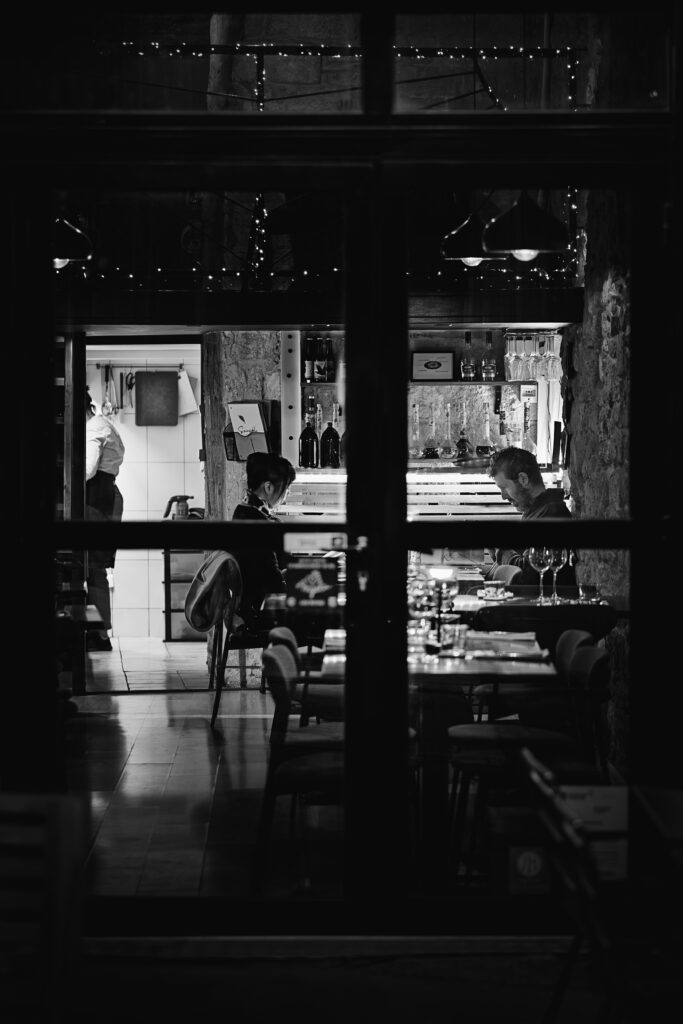 Photographie en noir et blanc de la devanture d'un restaurant. Au travers des vitres on peut apercevoir un couple, l'homme regardant son téléphone tandis que sa compagne regarde la carte. Derrière eux un bar avec des bouteilles bien alignées. Le couple est métaphoriquement séparé par la chambranle de la porte et de la vitre en premier plan. Dans le fond à gauche on peut apercevoir un cuisinier affairé.