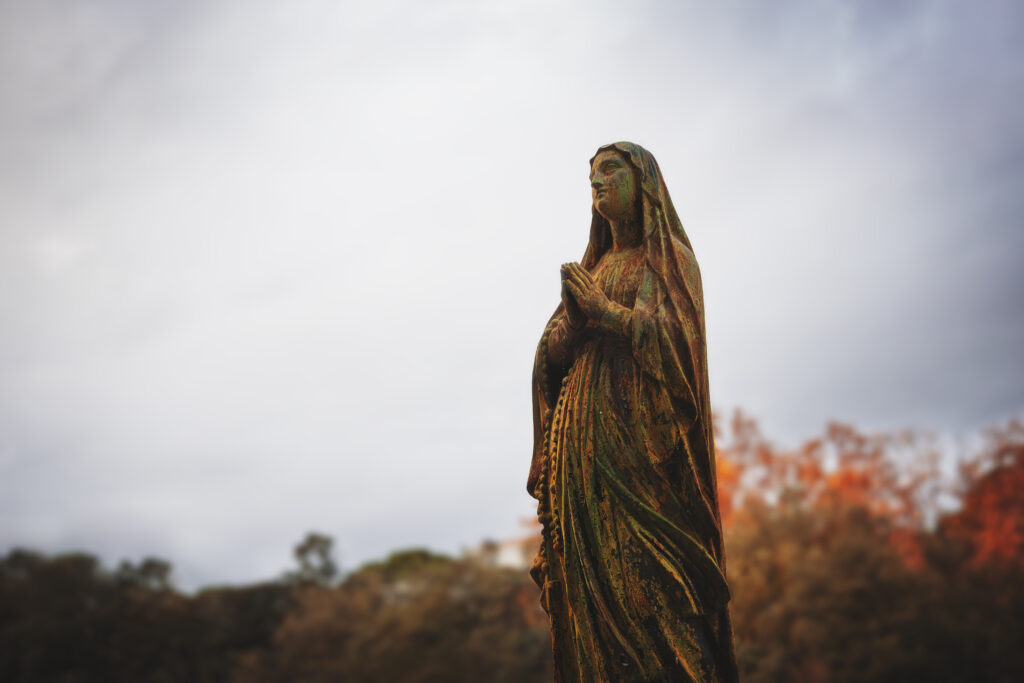 Photographie d'une statue de vierge en métal corrodé, donc la couleur rappelle celle des arbres rougis par l'automne.