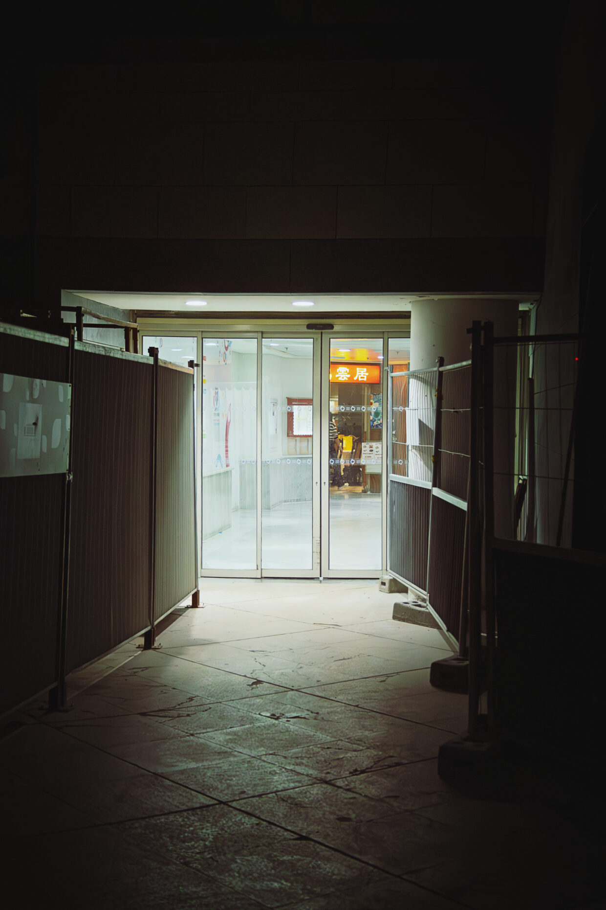 Photographie de rue, en couleur, montrant l'entrée d'un immeuble. La photographie est assez sombre, car prise en nocturne, seul le couloir du batiment photographié est éclairé, ainsi qu'une pancarte d'une restaurant asiatique dans le fond du couloir, face à l'entrée. De part et d'autre de cette entrée des panneaux de chantiers sont disposés.