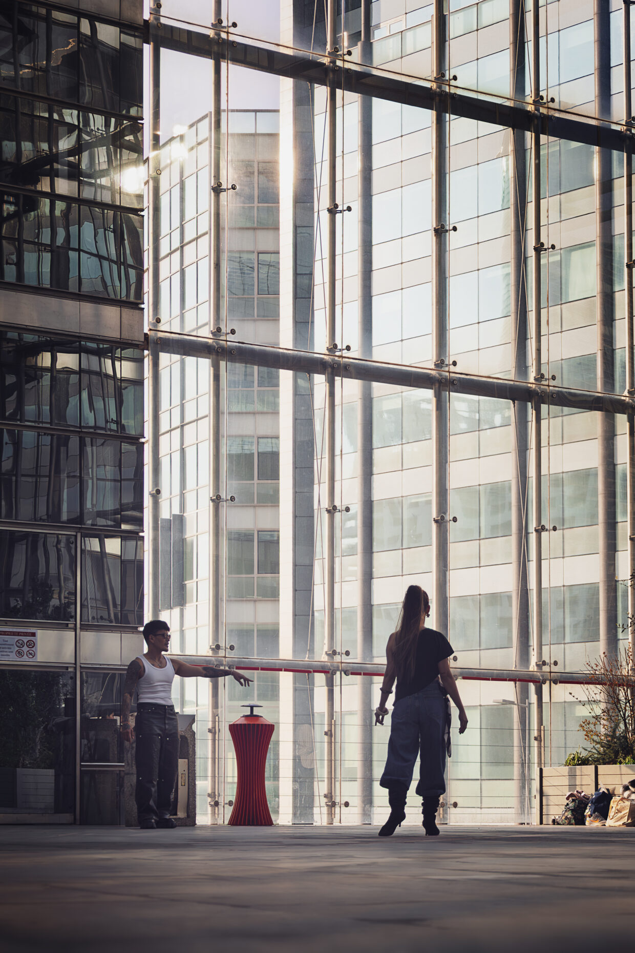 Photographie de rue, à l'intérieur d'un batiment de la Défense à Paris. Deux personnes se font face, vers le bas de la photographie, l'homme tendant le bras et semblant désigner un endroit où la femme, en talon haut et tenue décontractée doit aller. Derrière eux, une large baie vitrée sur toute la hauteur de la photographie, laisse apercevoir d'autres bâtiments tout aussi hauts.