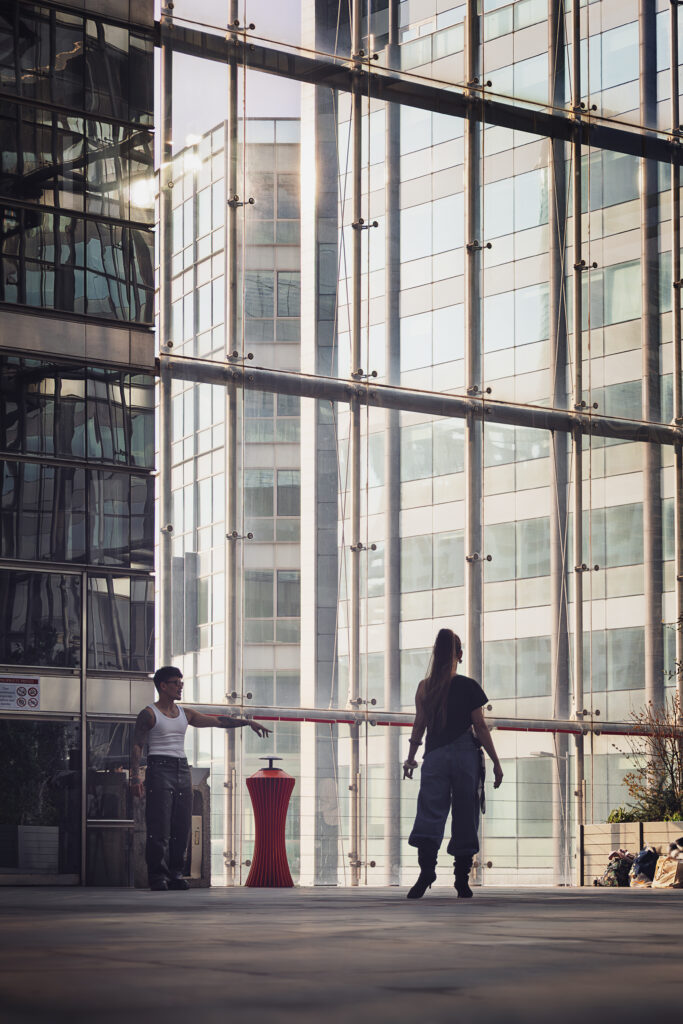 Photographie de rue, à l'intérieur d'un batiment de la Défense à Paris. Deux personnes se font face, vers le bas de la photographie, l'homme tendant le bras et semblant désigner un endroit où la femme, en talon haut et tenue décontractée doit aller. Derrière eux, une large baie vitrée sur toute la hauteur de la photographie, laisse apercevoir d'autres bâtiments tout aussi hauts.