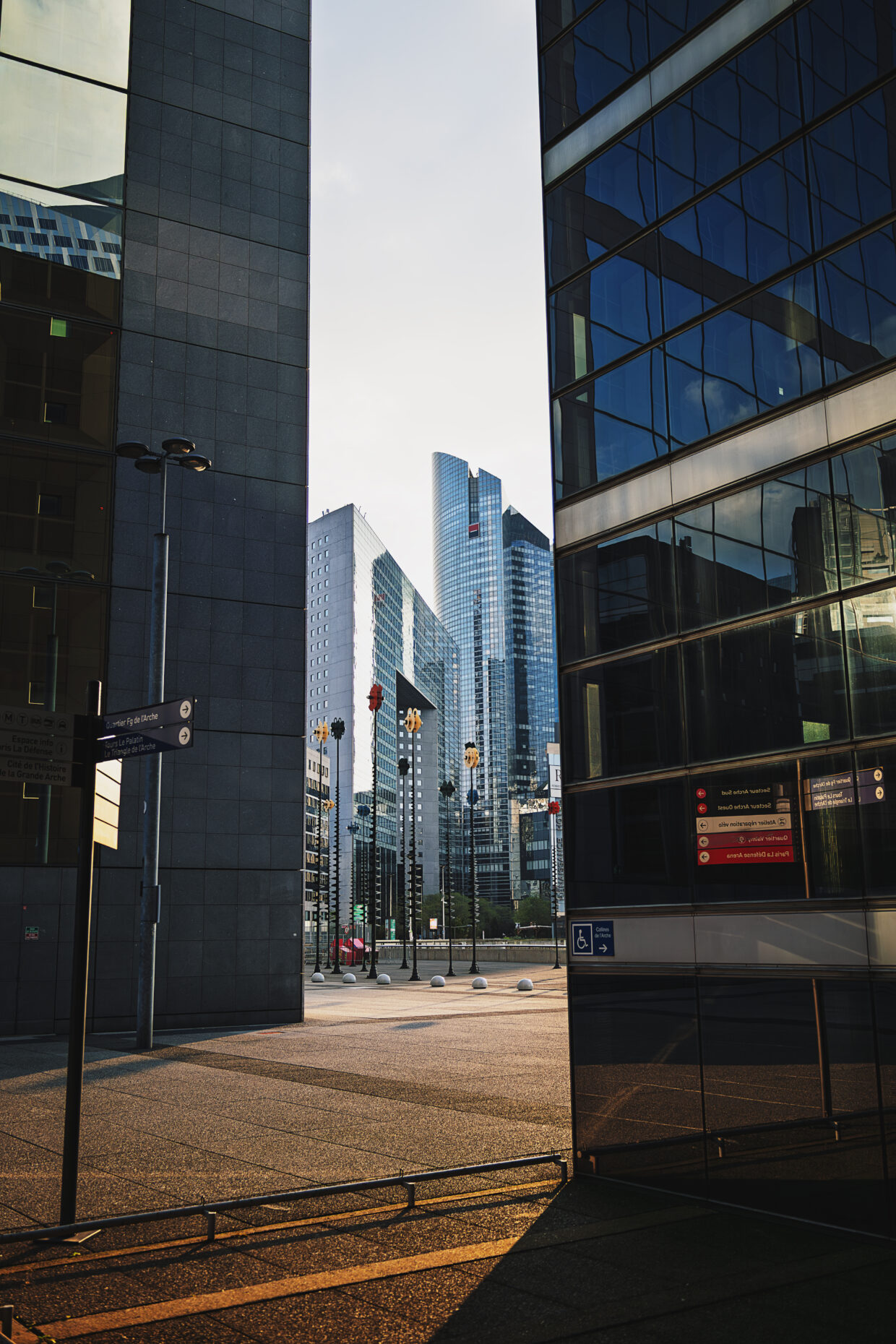 Photographie en couleur de plusieurs bâtiments de la Défense à Paris. De part et d'autre de la photographie, deux buildings très proches encadre une fenêtre artificielle laissant voir au loin d'autres bâtiments plus lointains. La lumière de la fin de journée est dorée, presque rasante, donnant un contraste important entre le verre bleuté des tours et le goudron rehaussé de dorure du sol.
