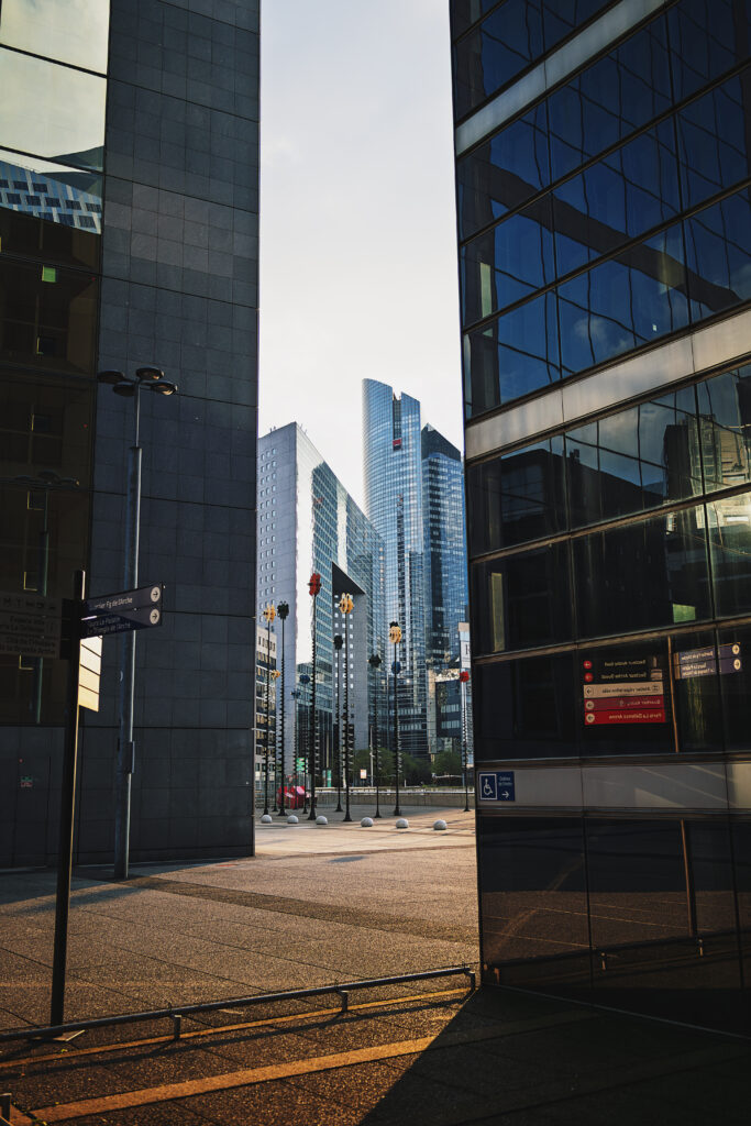 Photographie en couleur de plusieurs bâtiments de la Défense à Paris. De part et d'autre de la photographie, deux buildings très proches encadre une fenêtre artificielle laissant voir au loin d'autres bâtiments plus lointains. La lumière de la fin de journée est dorée, presque rasante, donnant un contraste important entre le verre bleuté des tours et le goudron rehaussé de dorure du sol.