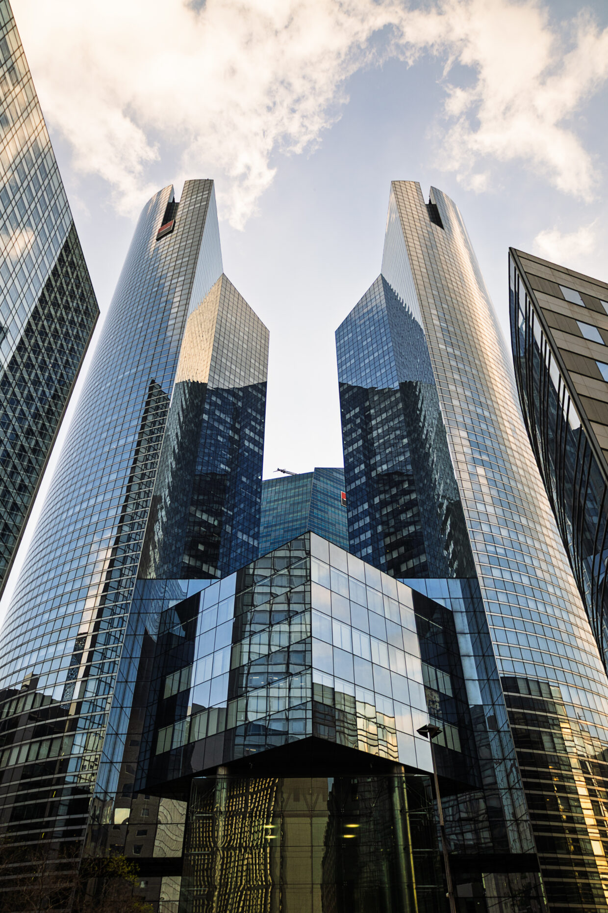 Photographie en couleur montrant la tour de la Société Générale à la Défense à Paris. La photographie se veut symétrique, centrée sur le vide entre les deux tours de la banque. Les façades de verre reflètent de nombreux bâtiments aux alentours, parfois très déformés par l'orientation des fenêtres.