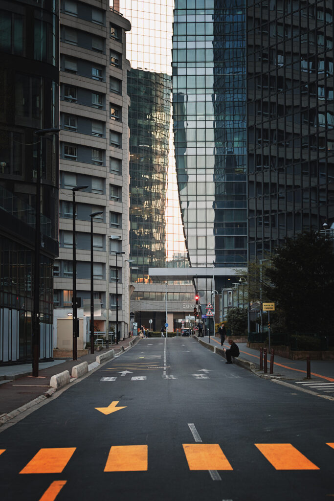 Photographie de rue, en couleur, montrant une route au milieu de grands immeubles, à la Défense à Paris. Se font face des bâtiments de logements, à gauche, et d'autres de bureaux, à droite. La rue est quasiment déserte, aucun véhicule n'est visible, et une personne est assise sur le trottoir, en train de regarder son téléphone. Au premier plan, en jeune orangé, un passage piéton temporaire attire l'oeil.