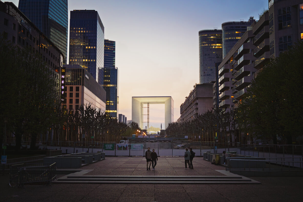 Photographie de rue en couleur montrant l'arche de la Défense à Paris, centrée, au crépuscule. De part et d'autre de l'arche de nombreux bâtiments se reflètent les uns les autres, grâce à leurs immenses façades de verre. Plusieurs fenêtres sont allumées ça et là. En bas, sur la promenade, plusieurs personnes se baladent.