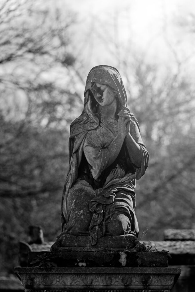 Photographie en noir et blanc d'une statue de femme, sur une tombe, à genoux, les mains jointes près de son épaule, le regard baissé vers la gauche du cliché. Derrière elle, en fond, plusieurs branches d'arbres et d'arbustes sont visible.