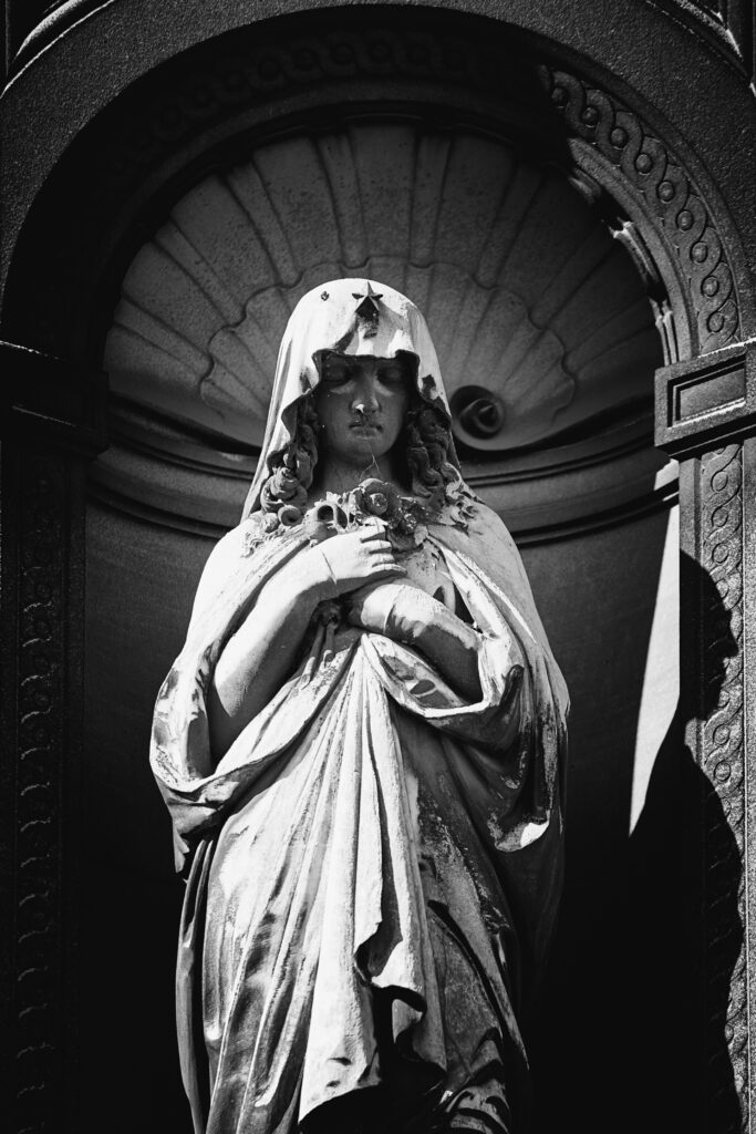 Photographie en noir et blanc d'une statue dans une alcôve. Elle représente une femme, voilée, dont les mains sont repliées sur la poitrine, tenant un bouquet de fleur. Sur le voile, au niveau du front, une étoile à cinq branche est visible. La poussière s'est accumulée sur la statue, masquant certains de ses traits, et rehaussant le drapé de ses vêtements.