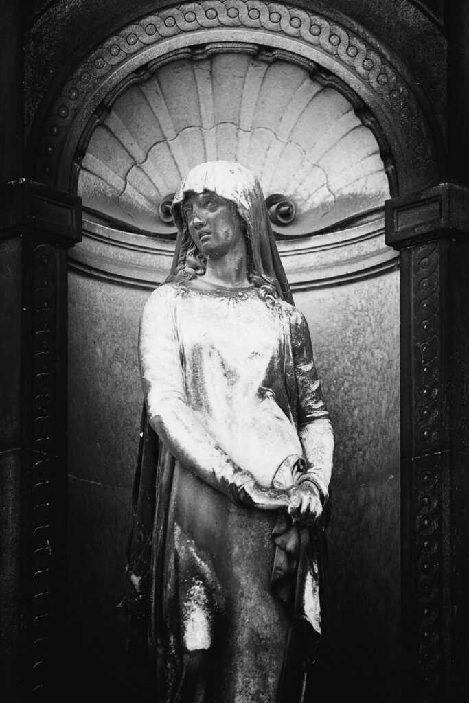 Photographie en noir et blanc d'une statue dans une alcôve. Elle représente une femme, voilée, dont les traits sont masqués par la poussière et la végétation des dernières années. Sa tête est tournée vers la gauche, ses lèvres légèrement entrouvertes. Ses mains sont jointes sur sa hanche droite.