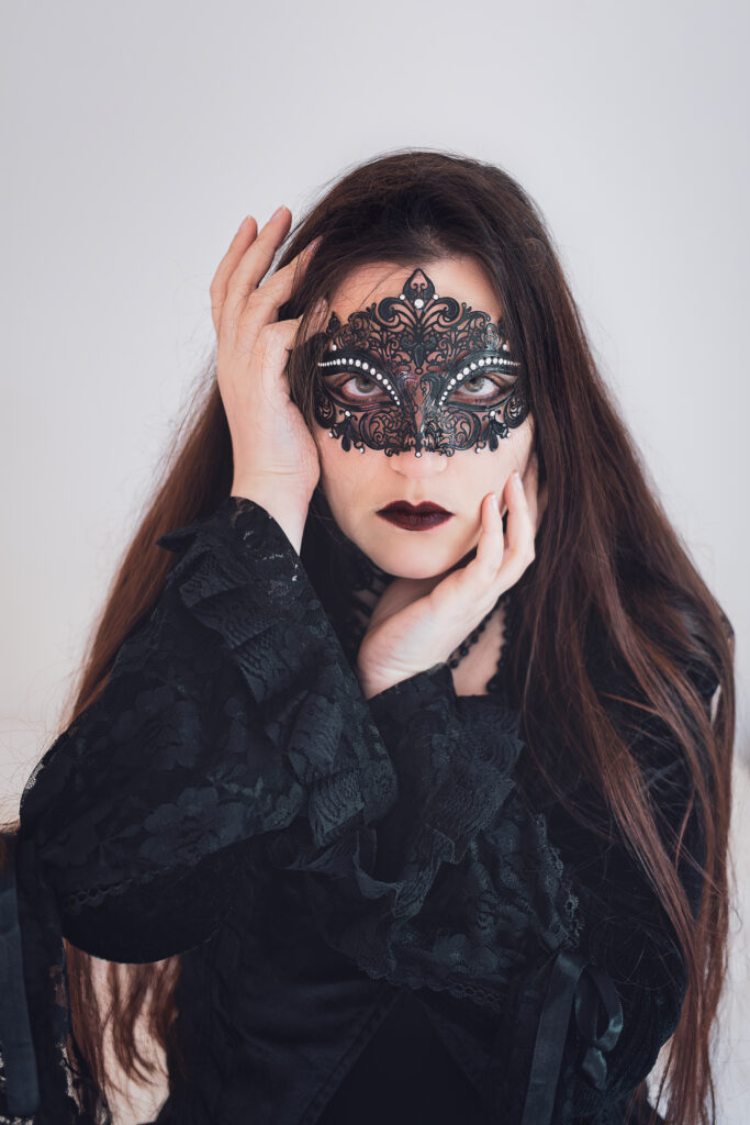 Une personne aux cheveux longs et habillée en noir fixe l'objectif. Elle est masquée avec un loup en fer, comme de la dentelle noire. Ses mains entourent son visage. Elle est maquillée de façon assez sombre, autant aux yeux qu'aux lèvres.