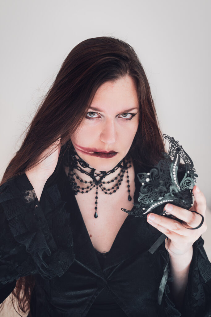 Une personne aux cheveux longs et habillée en noir fixe l'objectif. Elle tient dans sa main gauche un masque. Sa main droite est cachée par ses cheveux, proche de son visage. Son regard est fixe, elle semble en colère. Son rouge à lèvre est à moitié effacé, une longue trace est visible sur sa joue droite.