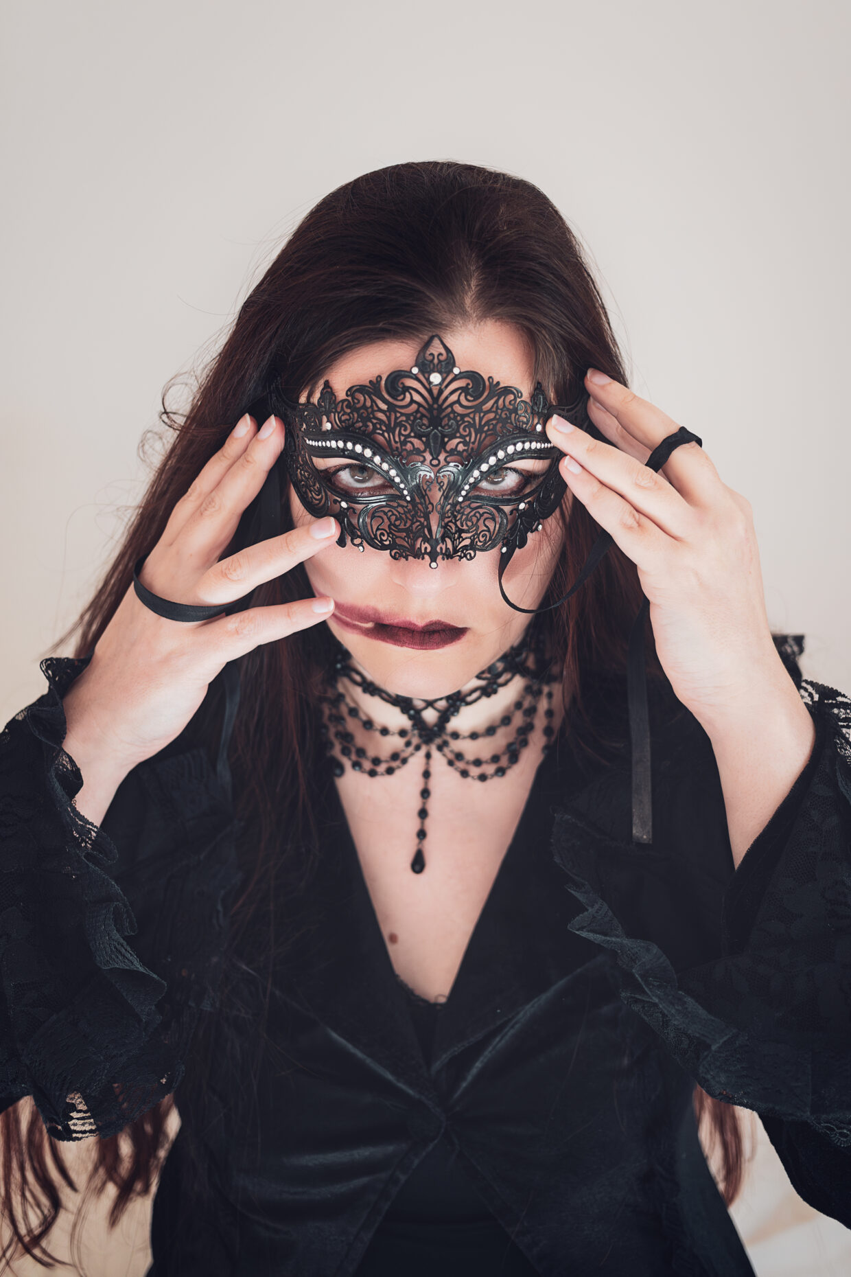 Une personne aux cheveux longs et habillée en noir fixe l'objectif. Elle est masquée avec un loup en fer, comme de la dentelle noire. Elle tient le masque elle même sur son visage. Son regard trahit de la colère. Son rouge à lèvre est à moitié effacé, une longue trace est visible sur sa joue droite.