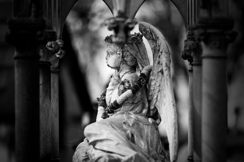 Photographie en noir et blanc d'une sculpture d'ange au cimetière du Père Lachaise. L'ange est un enfant et la statue est très abimée, le bas de son visage, vu de profil ayant disparu.