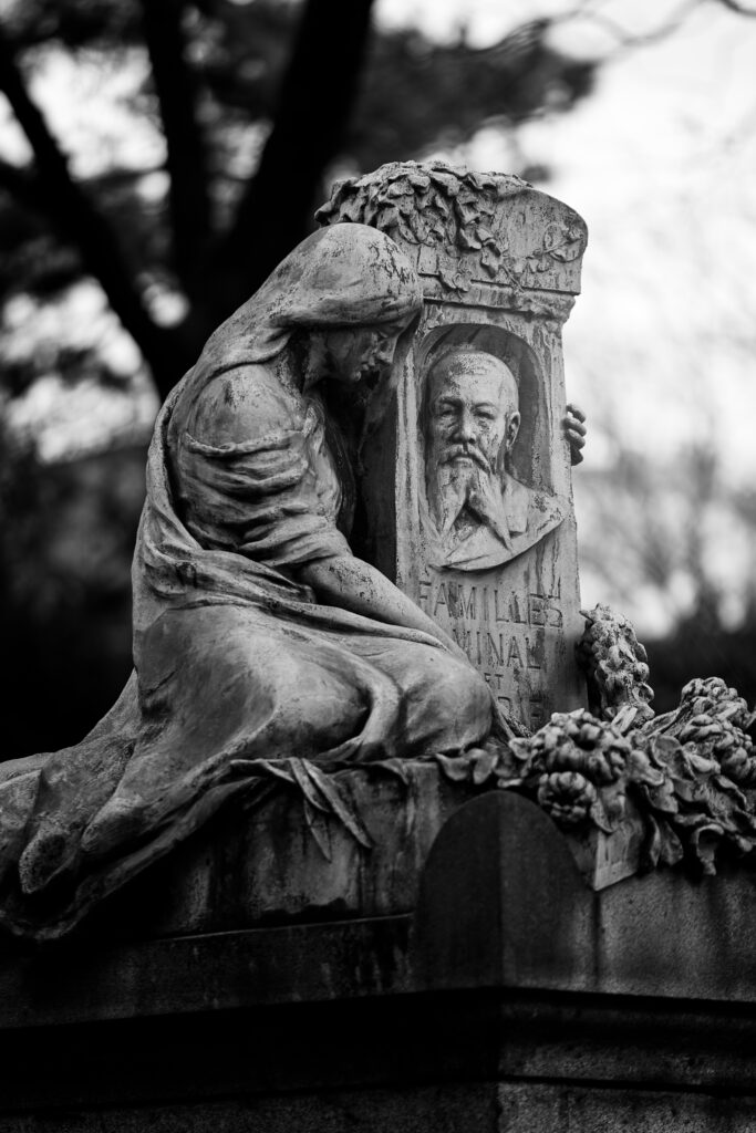 Photographie en noir et blanc d'une sculpture sur une tombe. Elle représente une femme dans des habits très amples, voilée, tenant une pierre tombale sur laquelle la tête d'un homme barbu est représentée.