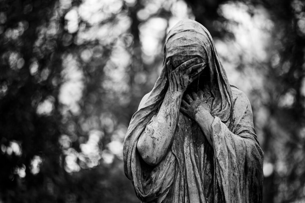 Photographie en noir et blanc d'une statue, probablement féminine, entièrement drapée, qui est dans une position de pleurs. L'une de ses mains cachant son visage. Dans le fond les branches des arbres sont visibles dans un bokeh doux.