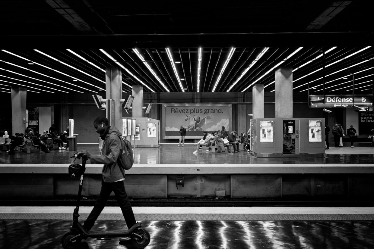 Photographie en noir et blanc dans le métro parisien, station La Défense. Au premier plan un homme part vers la gauche avec une trottinette. Au second plan de nombreuses personnes sont assises, la plupart en train de regarder leur téléphone. Les lignes de fuite amène vers une publicité d'une personne sur une licorne ailée, sur titrée 'Rêvez plus grand".
