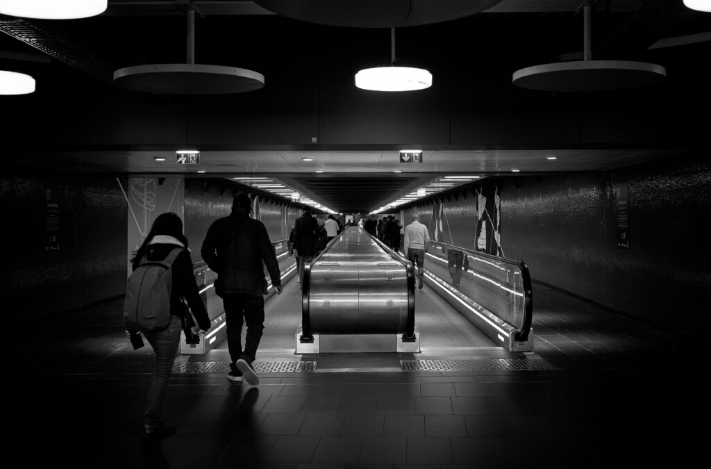 Photographie en noir et blanc du métro parisien, montrant avec un point de fuite vers le centre, plusieurs personnes prenant des tapis roulants. Sur le haut de la photographie plusieurs lampadaires modernes ronds sont visibles.