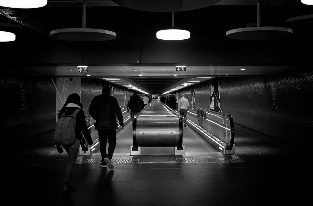 Photographie en noir et blanc du métro parisien, montrant avec un point de fuite vers le centre, plusieurs personnes prenant des tapis roulants. Sur le haut de la photographie plusieurs lampadaires modernes ronds sont visibles.