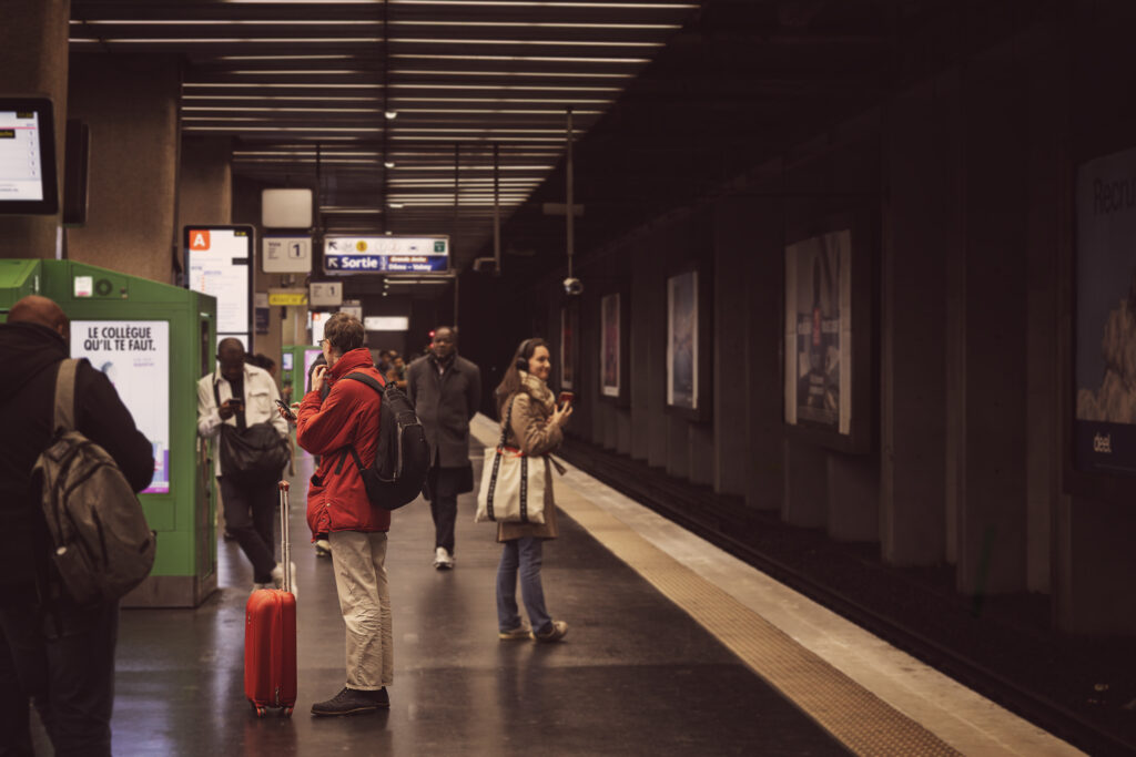 Photographie en couleur d'une scène dans le métro parisien, station La Défense, où des voyageurs attendent le train. La mise au point est faite sur un homme, dont le manteau et la valise sont de la même couleur : un rouge tirant légèrement vers le orange.
