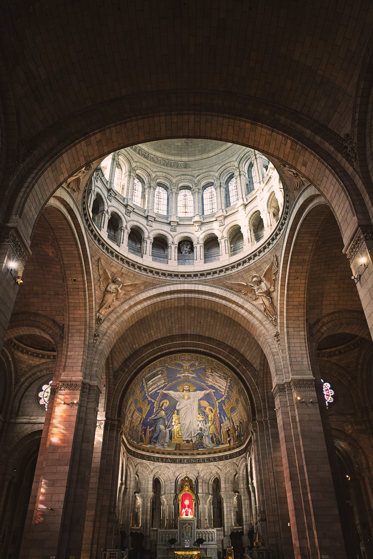 Photographie en couleur de la coupole du Sacré-Coeur à Paris. Au centre, en haut, la coupole soutenue par des voutes crée un puits de lumière éclairant le reste de la photographie. Au centre, en dessous de la coupole, quatre anges tenant chacun dans leurs main les instruments de la Passion du Christ. Au centre se situe la grande mosaïque montrant le Christ, bras ouverts, divers figures historiques dont Jeanne d'Arc, et personnages ayant contribué à l'histoire Chrétienne ou de la basilique.