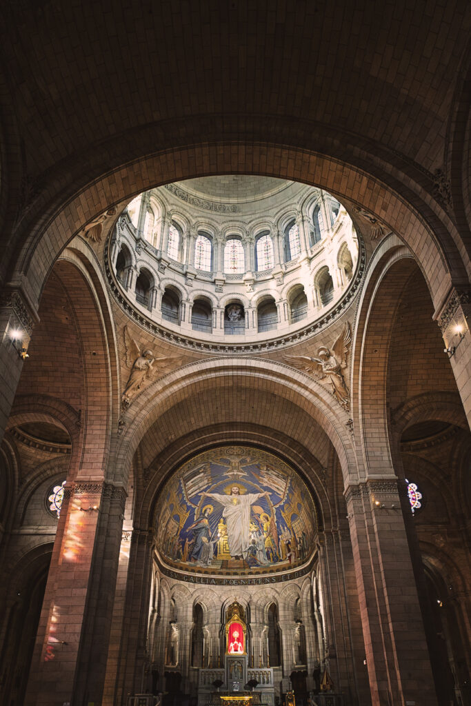Photographie en couleur de la coupole du Sacré-Coeur à Paris. Au centre, en haut, la coupole soutenue par des voutes crée un puits de lumière éclairant le reste de la photographie. Au centre, en dessous de la coupole, quatre anges tenant chacun dans leurs main les instruments de la Passion du Christ. Au centre se situe la grande mosaïque montrant le Christ, bras ouverts, divers figures historiques dont Jeanne d'Arc, et personnages ayant contribué à l'histoire Chrétienne ou de la basilique.
